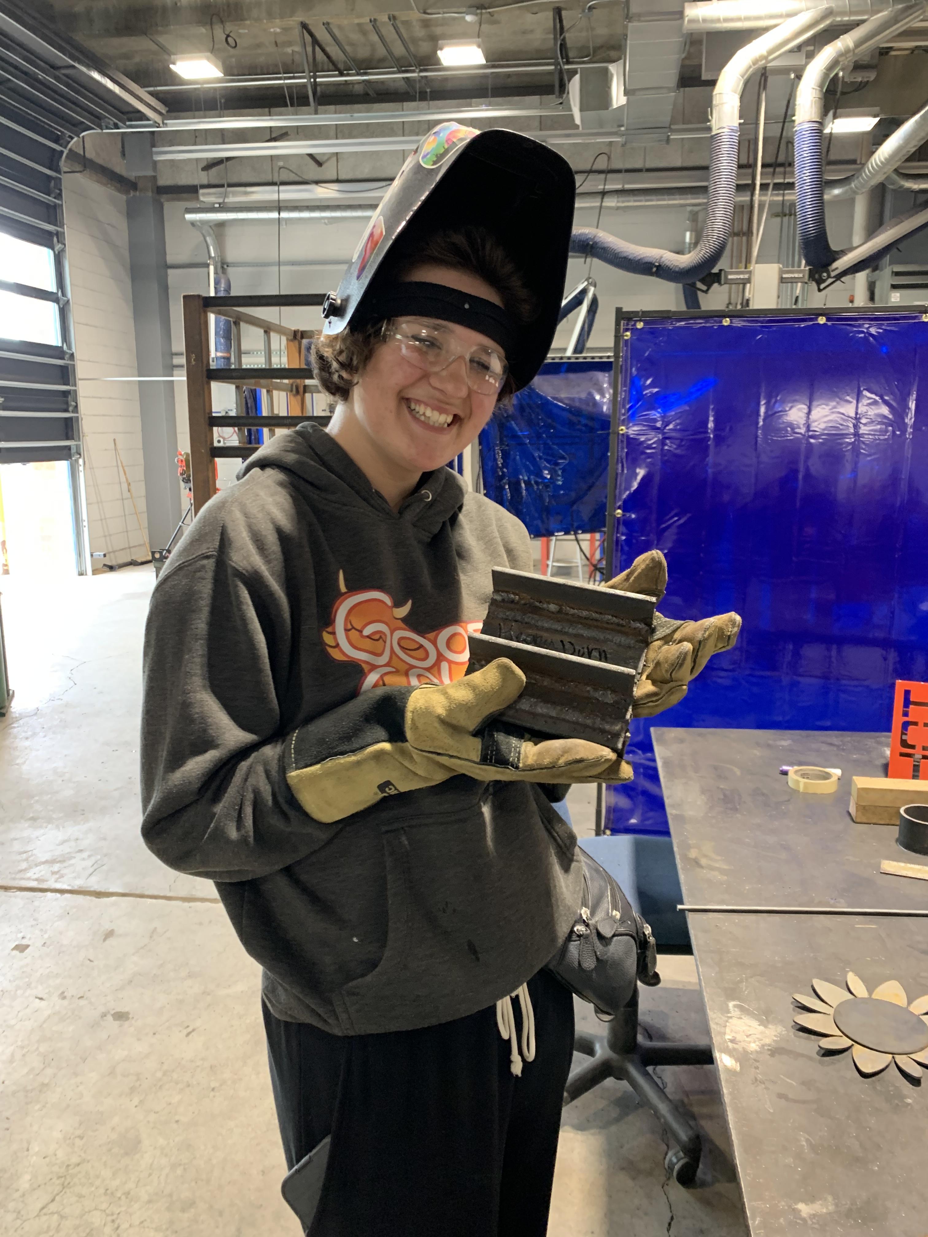 Keona in a welding mask and gloves holding her project. 